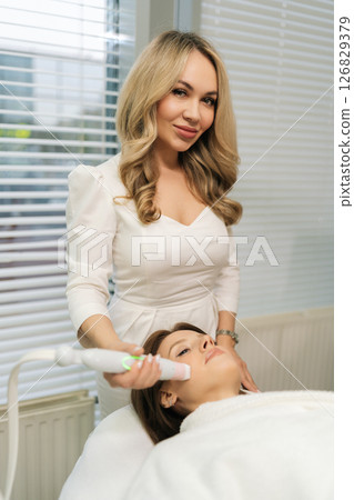 Vertical portrait of positive beautician performing mesotherapy procedure looking at camera, targeting facial skin rejuvenation with precision microinjection techniques in clinic of aesthetic medicine Vertical portrait of positive beautician performing mesotherapy procedure looking at camera, targeting facial skin rejuvenation with precision microinjection techniques in clinic of aesthetic medicine 126829379
