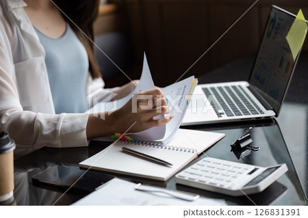 Diligent Asian woman reviewing documents and notes while working on a laptop in a modern office. 126831391