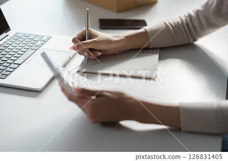 Diverse businesswoman taking notes and organizing tasks while working on her laptop, highlighting productivity in a home office. 126831405