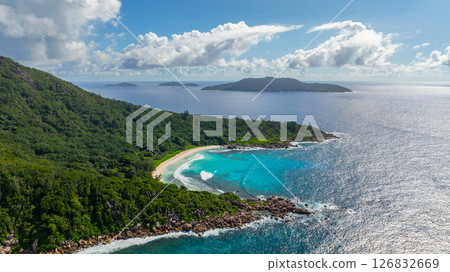 Waves gently roll onto a white sandy beach, bordered by lush green vegetation. La Digue, Seychelles. 126832669