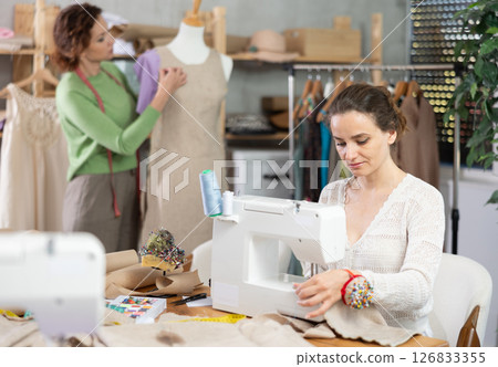 Woman tailor at work in cloth design shop. Companion work in background with mannequin Woman tailor at work in cloth design shop. Companion work in background with mannequin 126833355