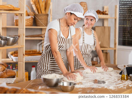 Two skilled female bakers in uniform kneading dough and rolling out dough into sausages before baking in bakehouse 126833384