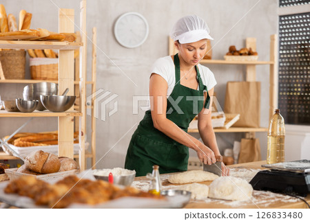 Middle-aged female baker cutting dough on table in bakehouse 126833480