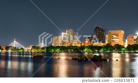 Panoramic night view of the Zakim Bridge and downtown Boston skyline reflecting over the Charles River with glowing city lights and trees lining the waterfront. Massachusetts, United States 126833671