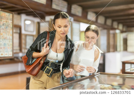 Young woman with teen daughter visitors stands near horizontal glass display case Young woman with teen daughter visitors stands near horizontal glass display case 126833701