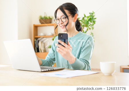 一位中年婦女一手拿著智慧型手機，一手在電腦上工作 126833776