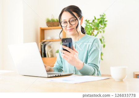 一位中年婦女一手拿著智慧型手機，一手在電腦上工作 126833778