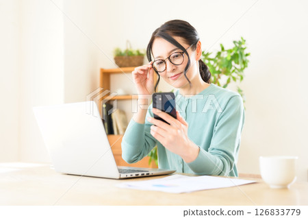 A middle-aged woman working on a computer with a smartphone in one hand A middle-aged woman working on a computer with a smartphone in one hand 126833779