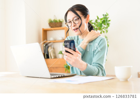 A middle-aged woman working on a computer with a smartphone in one hand A middle-aged woman working on a computer with a smartphone in one hand 126833781