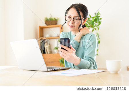 A middle-aged woman working on a computer with a smartphone in one hand 126833818