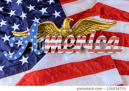 Striking golden eagle rests atop word America, set against waving American flag, symbolizing national pride. Striking golden eagle rests atop word America, set against waving American flag, symbolizing national pride. 126834733
