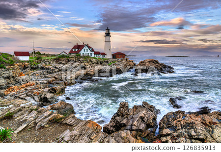 Portland Head Light stands prominently on the rocky shore of Cape Elizabeth, Maine, USA framed by dramatic ocean waves and colorful evening skies. The historic lighthouse and its red-roofed buildings 126835913