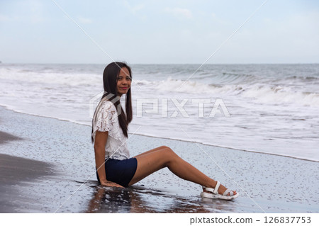 young woman sitting on wet black sand beach near waves 126837753