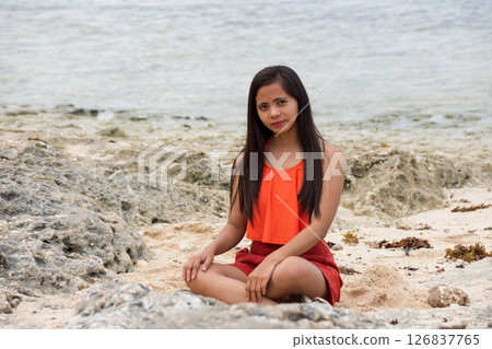 Young beautiful Asian woman in a swimsuit sits on a rocky beach by the sea, thoughtfully looking at the camera 126837765