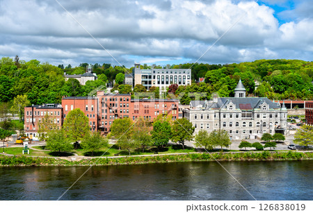 View across the Kennebec River toward historic downtown Augusta in Maine, USA, featuring Old Post Office, red-brick buildings and tree-covered hills under partly cloudy skies 126838019