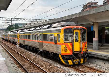 Taiwan Railways train stands at Ruifang Station on a cloudy day, with its colorful orange and yellow livery contrasting against the muted tones of the concrete platform Taiwan Railways train stands at Ruifang Station on a cloudy day, with its colorful orange and yellow livery contrasting against the muted tones of the concrete platform 126842493