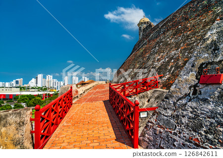 Sloped brick ramp with vivid red railings climbs alongside the weathered stone wall of Castillo San Felipe de Barajas in Cartagena, Colombia. The colonial fortification rises under a clear blue sky 126842611