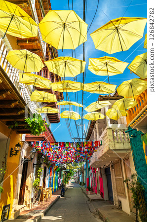 Bright yellow umbrellas and international flags hang above a narrow pedestrian street lined with colorful colonial buildings in Cartagena, Colombia. The lively decorations create a festive atmosphere 126842782