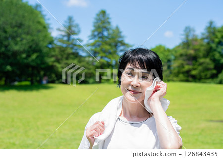 A middle-aged woman wiping off sweat with a towel after exercising in the park 126843435