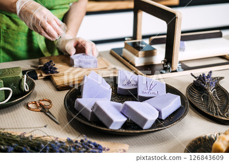Person stamps lavender soap bar in a home workshop with dried flowers and artisan tools. Sustainable goods, handmade skincare, eco-friendly production, zero waste soap Person stamps lavender soap bar in a home workshop with dried flowers and artisan tools. Sustainable goods, handmade skincare, eco-friendly production, zero waste soap 126843609