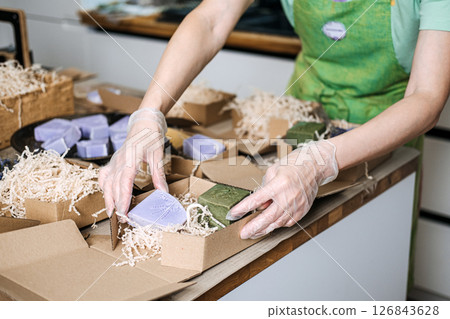 Person arranges lavender and olive oil soaps into kraft boxes with shredded filler at a worktable. Green business, sustainable packaging, soap making business, eco-friendly practices Person arranges lavender and olive oil soaps into kraft boxes with shredded filler at a worktable. Green business, sustainable packaging, soap making business, eco-friendly practices 126843628