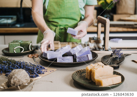 Person arranges freshly made lavender soap bars on a plate beside other handcrafted soaps and dried flowers. artisanal small business, low-waste packaging, wellness lifestyle 126843630