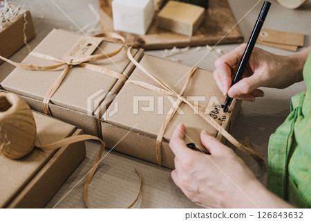 Person writes on a kraft tag attached to a ribbon-wrapped gift box near handmade soap. Handwritten tags, small business packaging, emotional branding, customer connection 126843632
