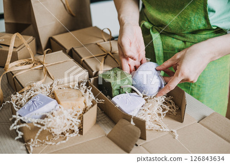 Person places handmade soaps and lavender sprigs into a kraft gift box with shredded filler. Artisanal skincare, eco-friendly packaging, handmade beauty gift, small batch soap 126843634