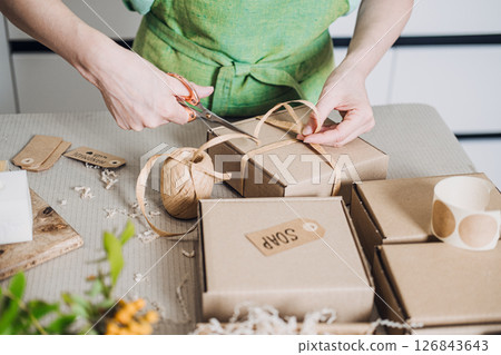 Person prepares ribbon-tied kraft boxes labeled soap in a minimal artisan workspace. Gen Z shopping habits, meaningful consumption, maker culture, personalized goods 126843643
