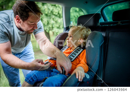 Toddler frowns while dad buckles him into car seat. Toddler frowns while dad buckles him into car seat. 126843984