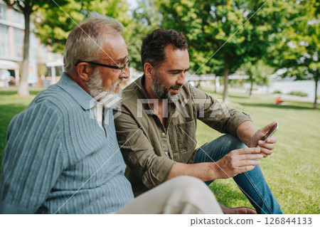 Son showing older father something in smartphone, sharing a joyful moment . 126844133