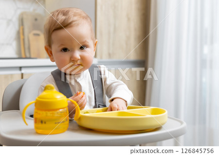Cute baby boy sitting on his chair and eating from his yellow plate 126845756
