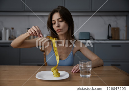 Skinny thin woman eating measuring tape demonstratively at home kitchen Skinny thin woman eating measuring tape demonstratively at home kitchen 126847203