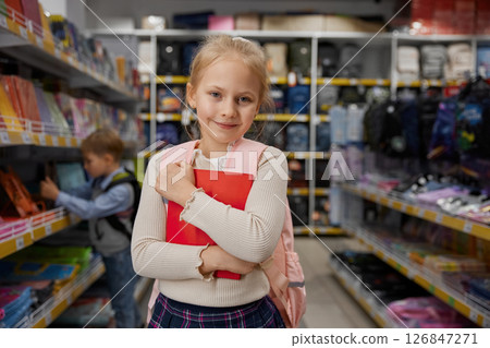Portrait of smiling girl child carrying red copybook in hands posing in shop Portrait of smiling girl child carrying red copybook in hands posing in shop 126847271