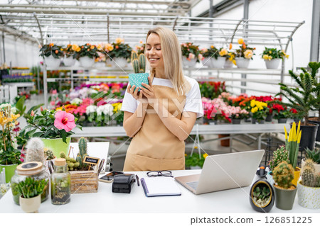 A smiling Caucasian woman, a florist, holding a cactus in her shop, next to a laptop and various plants for sale. 126851225