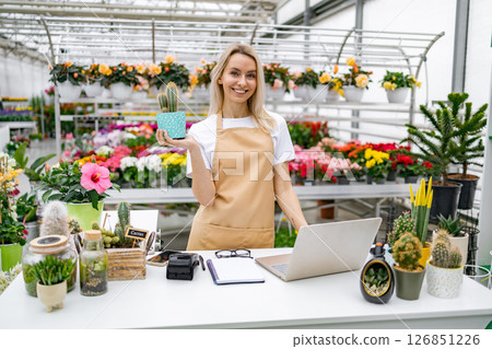 A Caucasian woman florist is smiling while holding a cactus in a shop filled with flowers. 126851226