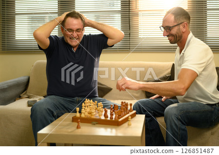 Elderly father and adult son playing a game of chess together in a living room. Father day concept Elderly father and adult son playing a game of chess together in a living room. Father day concept 126851487