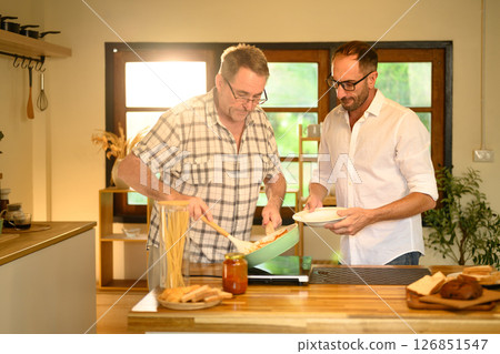 Happy senior man and grown son preparing pasta dish in a bright kitchen. Father day concept Happy senior man and grown son preparing pasta dish in a bright kitchen. Father day concept 126851547
