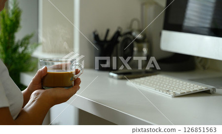 Close up of person holding a steaming cup of coffee while working in a home workspace 126851563