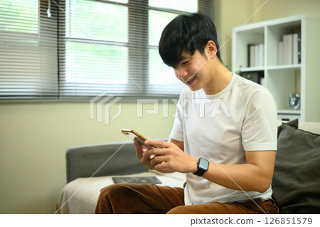 Smiling man using a smartphone while sitting in a relaxed living room 126851579