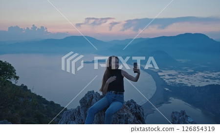 Happy beautiful young girl making a video or selfie holding a smartphone sharing moments of the trip using the app on a scenic viewpoint overlooking the mountains and sea Iztuzu beach at sunset Turkey Happy beautiful young girl making a video or selfie holding a smartphone sharing moments of the trip using the app on a scenic viewpoint overlooking the mountains and sea Iztuzu beach at sunset Turkey 126854319