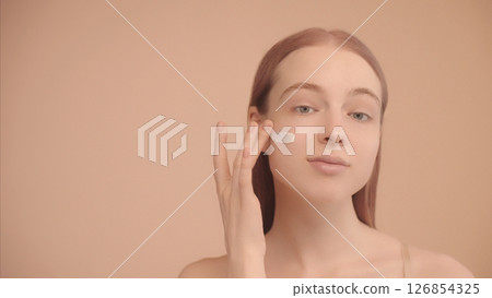 Portrait of a young Caucasian lady with smear of face cream on cheek Isolated Peach background. Woman applying moisturizing cream, looking at camera. Portrait of a young Caucasian lady with smear of face cream on cheek Isolated Peach background. Woman applying moisturizing cream, looking at camera. 126854325
