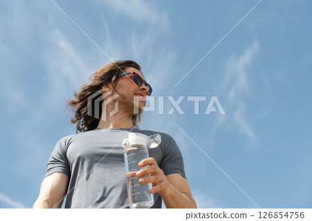 Man in athletic wear with water bottle outdoors against blue sky background. Summer heat weather. Hero view. Thirst. Close up. Banner. 126854756