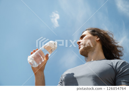 Handsome curly brunette man wear gray t-shirt with water bottle against blue sky background. Summer heat weather. Outdoors. Hero view. Close up. Banner. Handsome curly brunette man wear gray t-shirt with water bottle against blue sky background. Summer heat weather. Outdoors. Hero view. Close up. Banner. 126854764