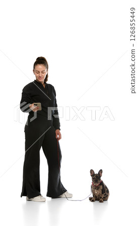 Calm French bulldog sitting by owner's feet on leash while woman smiles at phone, peaceful walk moment against white studio background. 126855449