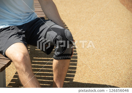 Man sitting on bench wearing knee brace to support painful leg. Athlete leg wearing knee support brace or elastic kneecap bandage. Sleeve for compression. Health problems and pain. 126856046