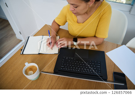 Focused Woman Planning Work Tasks While Writing in Notebook at Home Focused Woman Planning Work Tasks While Writing in Notebook at Home 126857066
