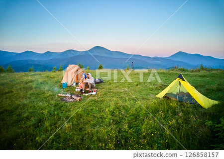 Scenic mountain campsite with two tents in morning. Two campers enjoying serene view of rolling mountains under clear sky. Picturesque sunrise landscape, creating tranquil and cozy outdoor experience. 126858157