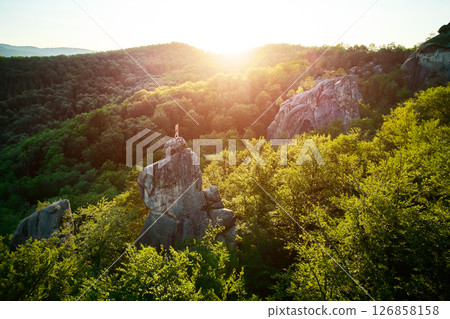 Aerial view of climber standing atop rock formation at Dovbush Rocks in Carpathian mountains, Ukraine. Sun sets, casting warm glow over lush, green forest, distant hills, creating breathtaking view. 126858158