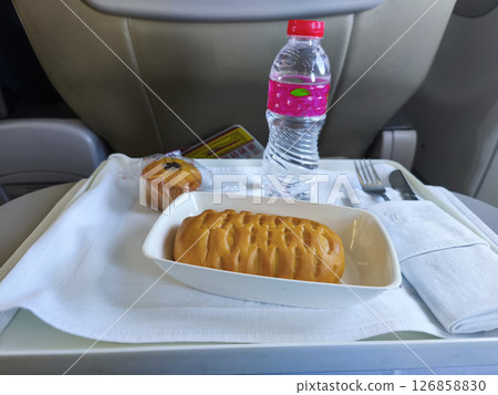 Airline meal with a water bottle and a muffin, viewed from a passenger's seat with an in-flight entertainment screen showing a colorful landscape. 126858830